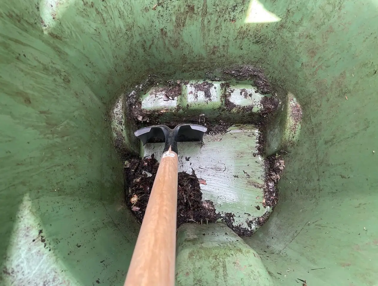 A person using a square-bladed transfer shovel to scrape stuck compost from the bottom of a green yard waste cart.